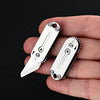 A hand holds a compact stainless steel pocket knife with a serrated blade and keychain attachment.