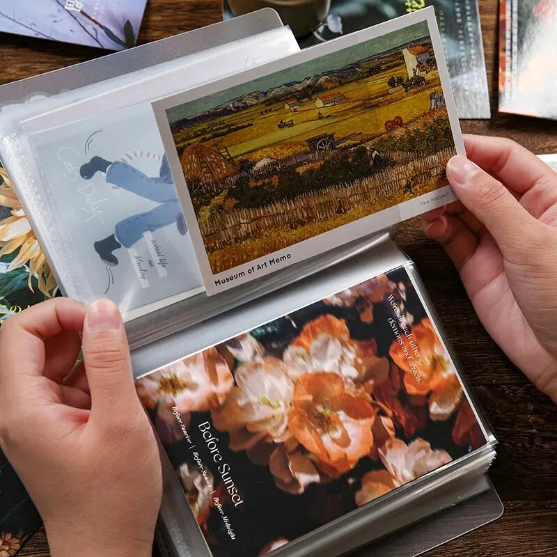 A hand holds an A6 photo storage booklet displaying a landscape painting and a close-up of flowers.