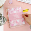 A pink envelope savings challenge planner with a gold spiral binding and a metal snap button is shown on a light pink surface