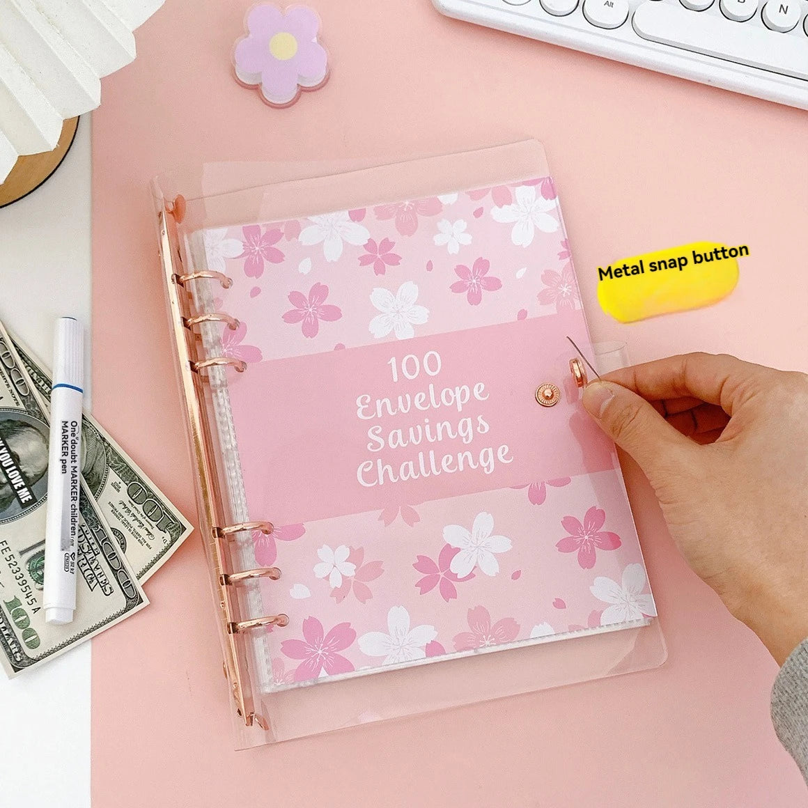 A pink envelope savings challenge planner with a gold spiral binding and a metal snap button is shown on a light pink surface