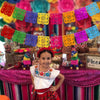 A young girl wearing a white shirt and red skirt stands in front of a colorful papel picado banner decoration.