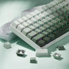 Vista cercana de un teclado mecánico blanco con teclas grises e iluminación RGB verde sobre una superficie clara.