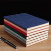 A stack of five premium A5 leather journals in shades of blue, pink, and green resting on a wooden surface.