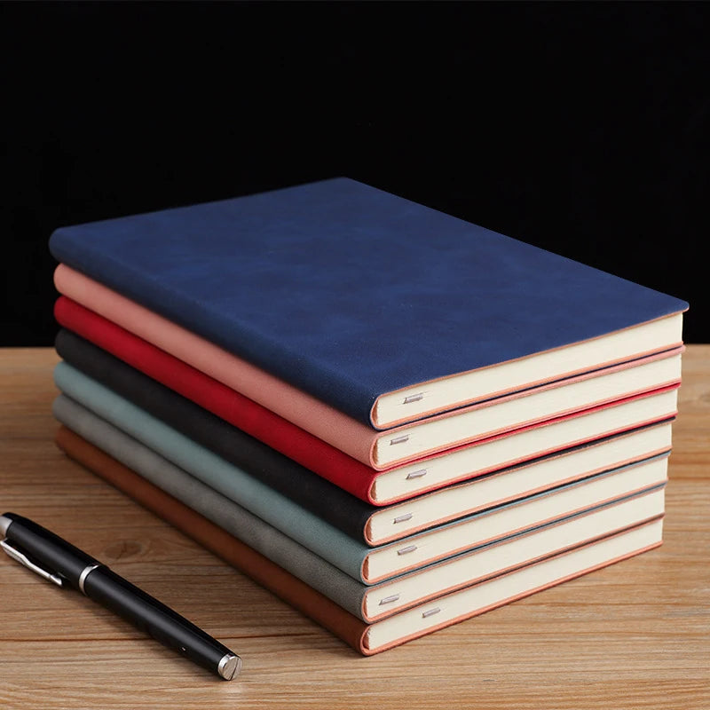 A stack of five premium A5 leather journals in shades of blue, pink, and green resting on a wooden surface.
