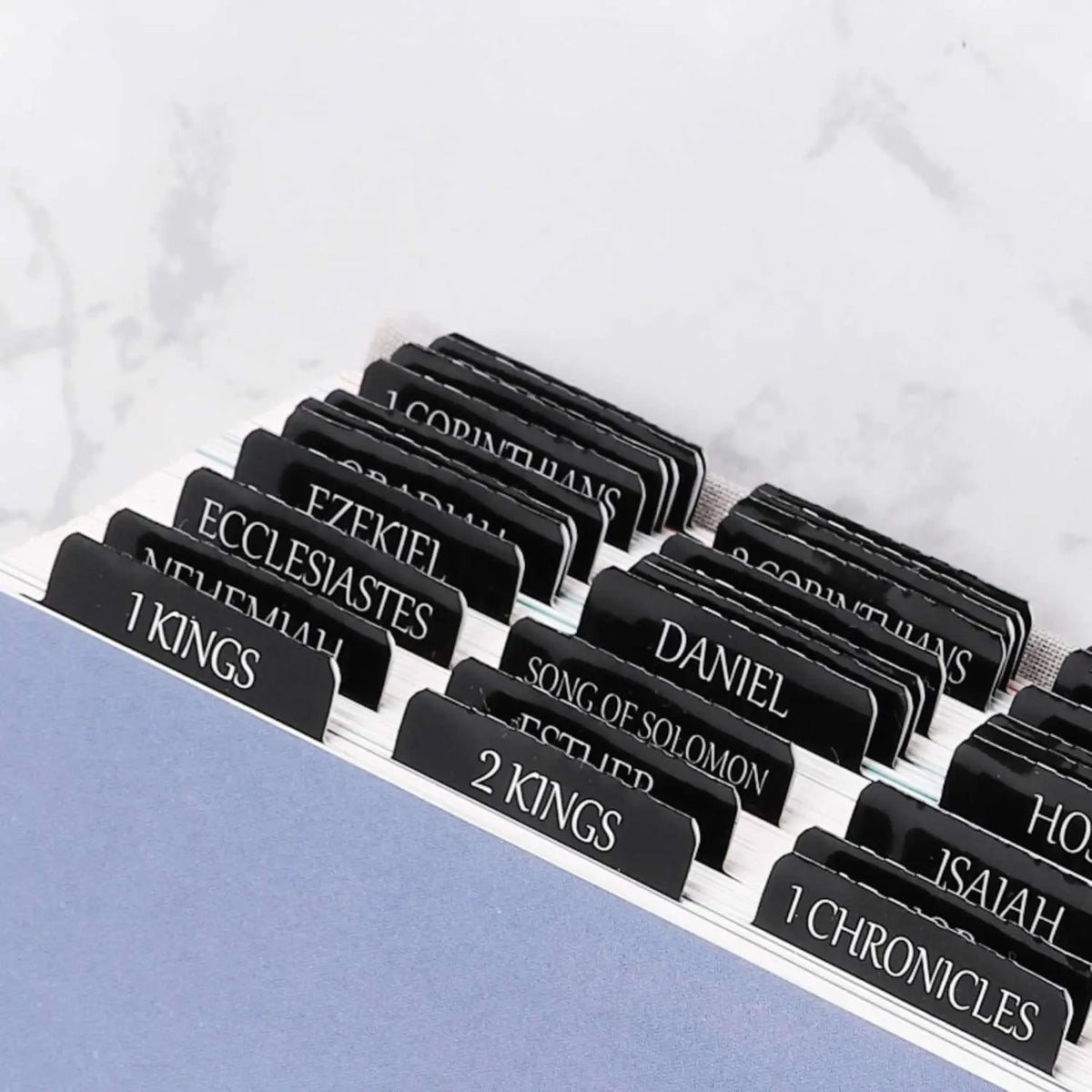 A collection of black laminated Bible tabs with biblical names and chapters laid out on a white marble surface.
