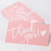 Pink thank you cards with white script text and heart accents stacked on a white surface