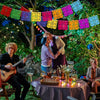 A family celebrating Día de Muertos with colorful papel picado banners strung between trees in an outdoor setting.