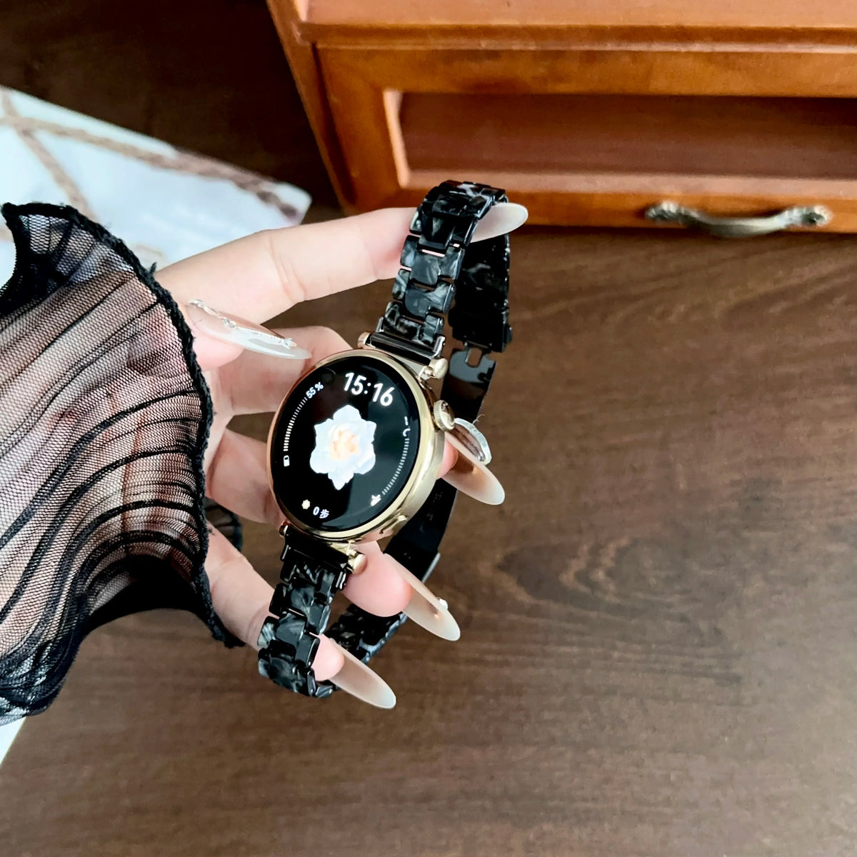 A hand wearing a black resin beaded watch with a gold-toned face and a black strap, displaying the time 15:16.
