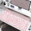 A large pink leopard print desk pad is displayed on a white desk next to a digital clock and monitor.