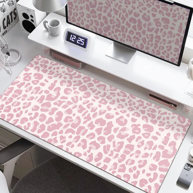 A large pink leopard print desk pad is displayed on a white desk next to a digital clock and monitor.