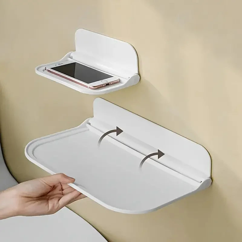 A hand is opening a white foldable wall shelf for no-drill storage, revealing a slot for a smartphone.