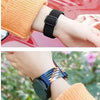 A close-up of a black braided loop watch band worn on a wrist next to an orange knitted sleeve.