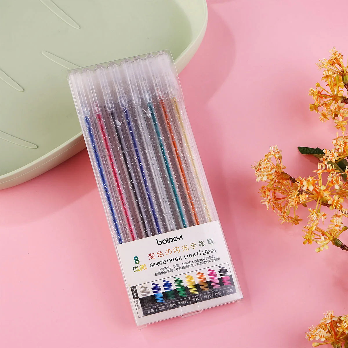 A clear plastic case containing a set of 8 colored glitter gel pens with tips, resting on a pink surface.