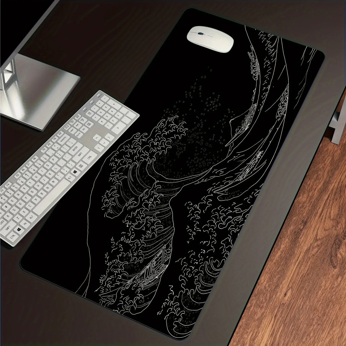 A black desk mouse pad featuring a white line drawing of a crashing wave and sea foam, with a white computer mouse resting on
