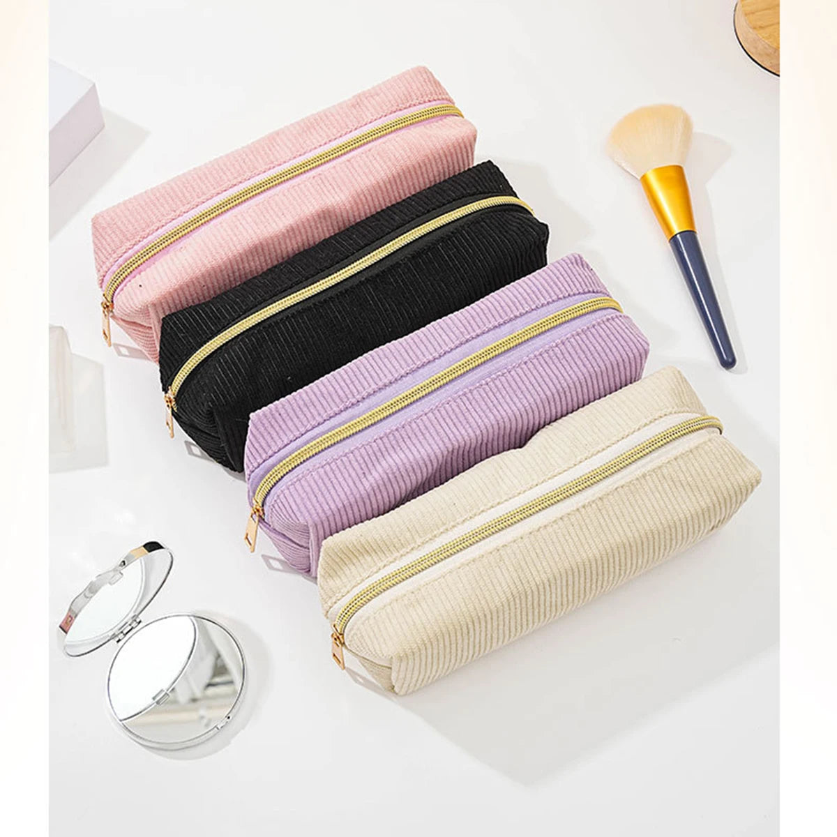 A collection of stacked pencil cases in pink, black, purple, and beige, with a gold-tipped pencil brush nearby.