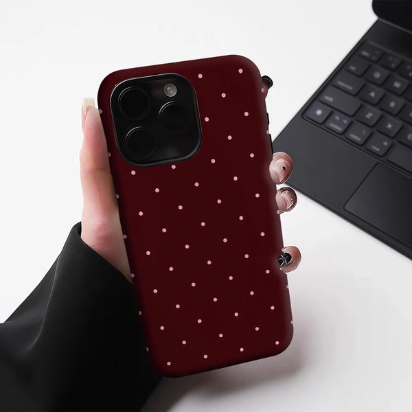 A hand holds a dark maroon phone case with small white polka dots, next to a black laptop.