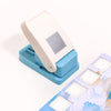 A white embossing punch machine with a light blue base and a sheet of patterned paper.