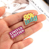A set of colorful enamel pins featuring an 80s baby design and a limited edition badge, held in a hand.