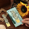 A hand holds a vintage artistic metal bookmark with a green tassel resting on a light blue paper against a dark wooden backgr