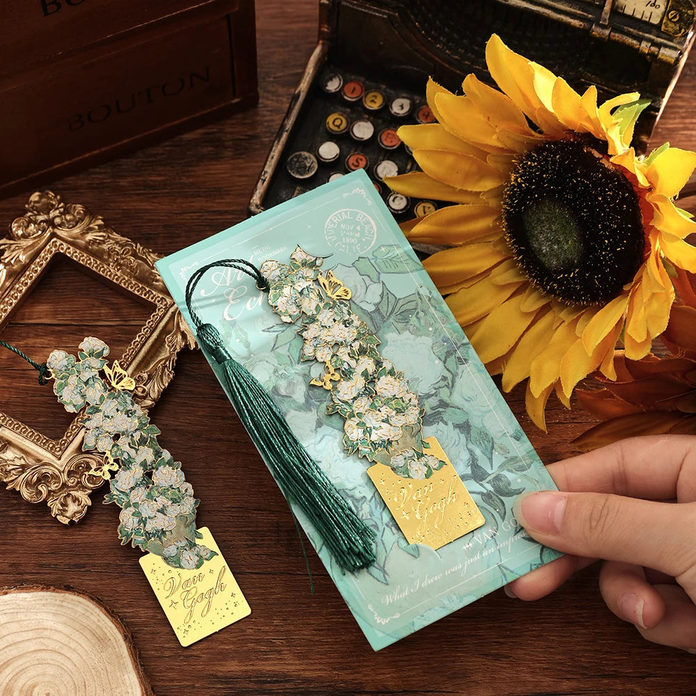 A hand holds a vintage artistic metal bookmark with a green tassel resting on a light blue paper against a dark wooden backgr