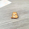 Broche de esmalte en forma de un gato atigrado naranja sentado sobre una superficie de madera.