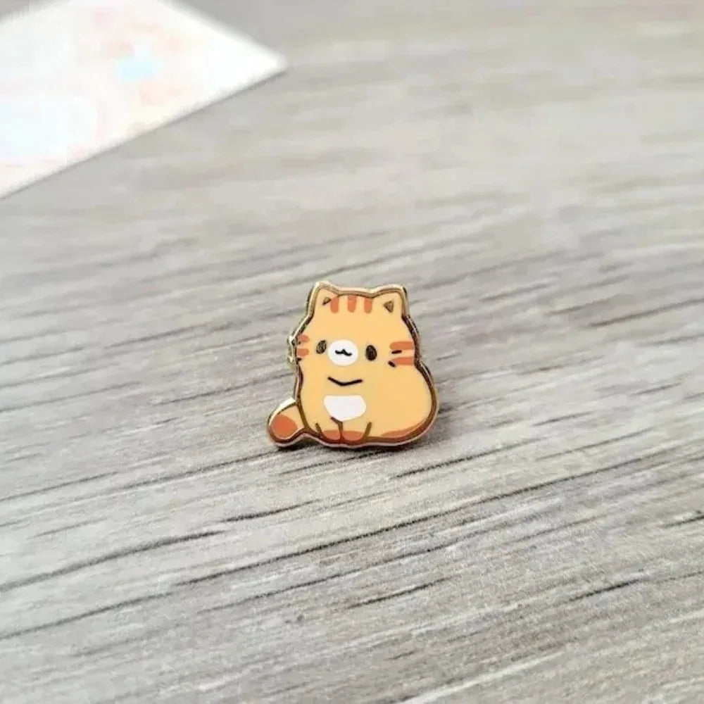 Broche de esmalte en forma de un gato atigrado naranja sentado sobre una superficie de madera.