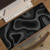 A black and gray abstract art desk mat with a non-slip rubber base is shown next to a laptop.