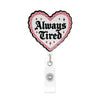 A heart-shaped badge reel with pink and white design featuring the text Always Tired