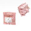 A pink linear mechanical keyboard switch is shown, featuring a white cross shape on the top housing.