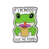 An enamel pin featuring a green frog with text that reads If I'm moody give me food