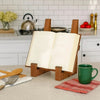A vintage wooden cookbook holder stands on a kitchen counter next to a green mug and a red cloth.
