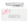 A large gray PU leather desk mat is shown with a laptop and keyboard placed on it, surrounded by pink cherry blossoms.