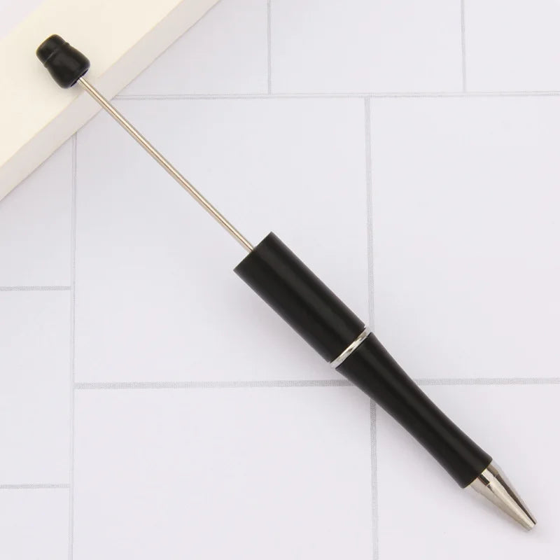 A black DIY beadable pen with a gold tip and a long metal shaft is shown on a white tiled surface.