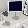 A white adjustable desktop stand holding a dark tablet and a heart-shaped phone.