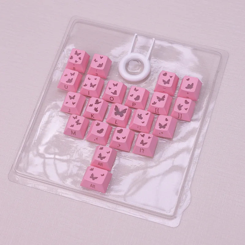 Eine Sammlung rosa Schmetterlings-Keycaps und ein weißer Ring in einer klaren Kunststoffschale.