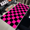 A pink and black checkered desk mat is displayed on a light wood surface next to a keyboard and gaming controller.