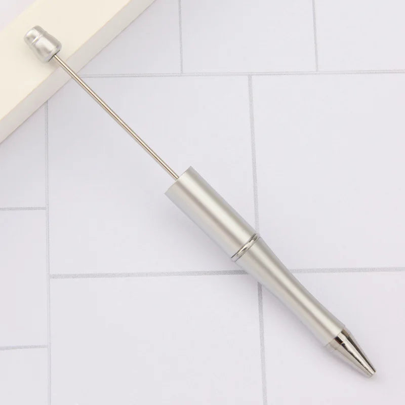 A silver DIY beadable pen with a pointed tip rests on a white tiled surface.