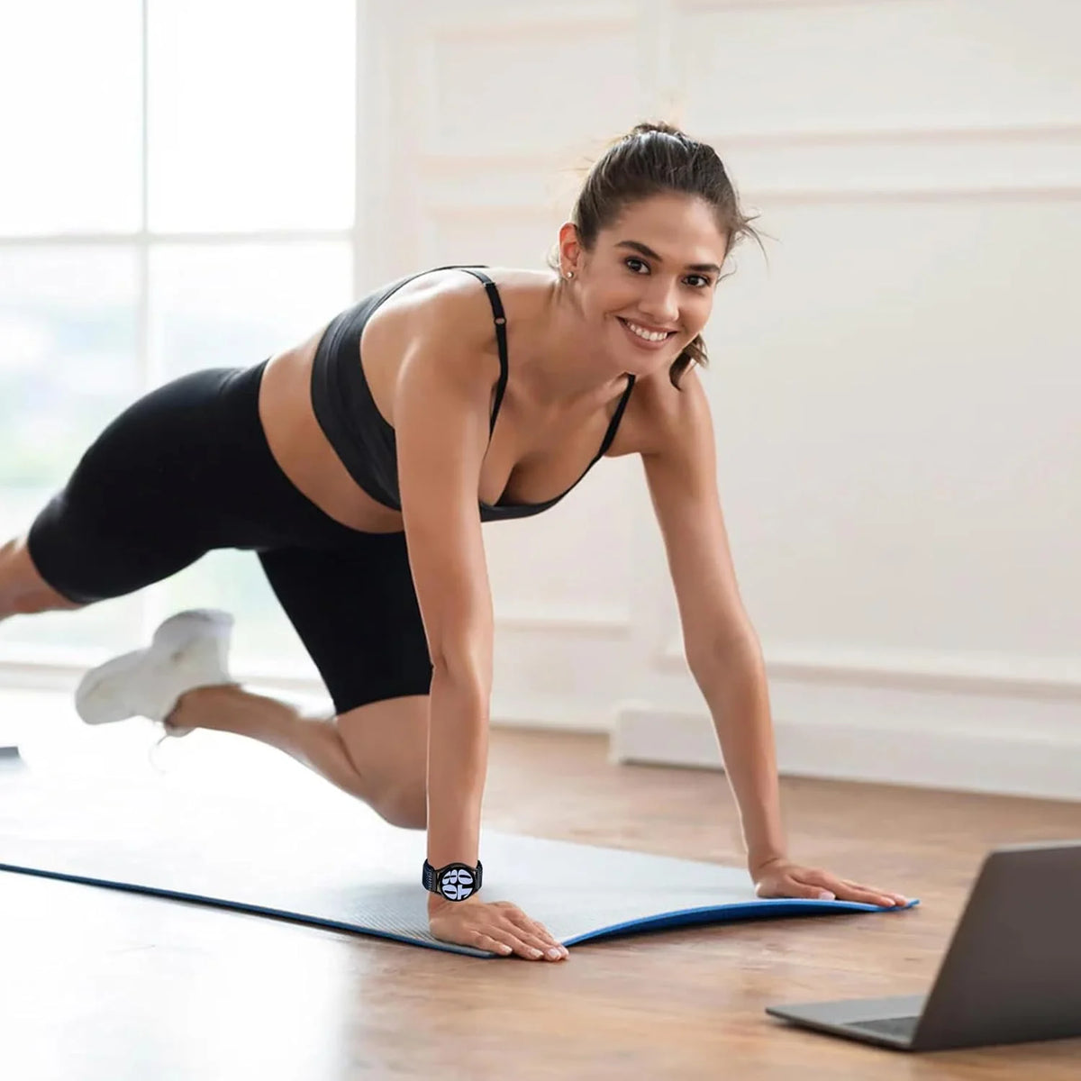 A woman in black athletic wear is performing a stretch on a yoga mat with a laptop nearby.