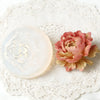 A pink rose-shaped wax melt and a gold-dusted flower are displayed on a white lace surface.