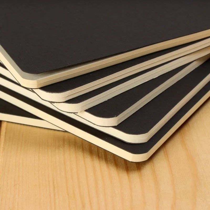 A stack of black A6 blank sketchbooks with light-colored covers resting on a wooden surface.