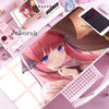 A close-up of a pink-haired anime girl resting on a purple desk mat with various accessories and a tablet.