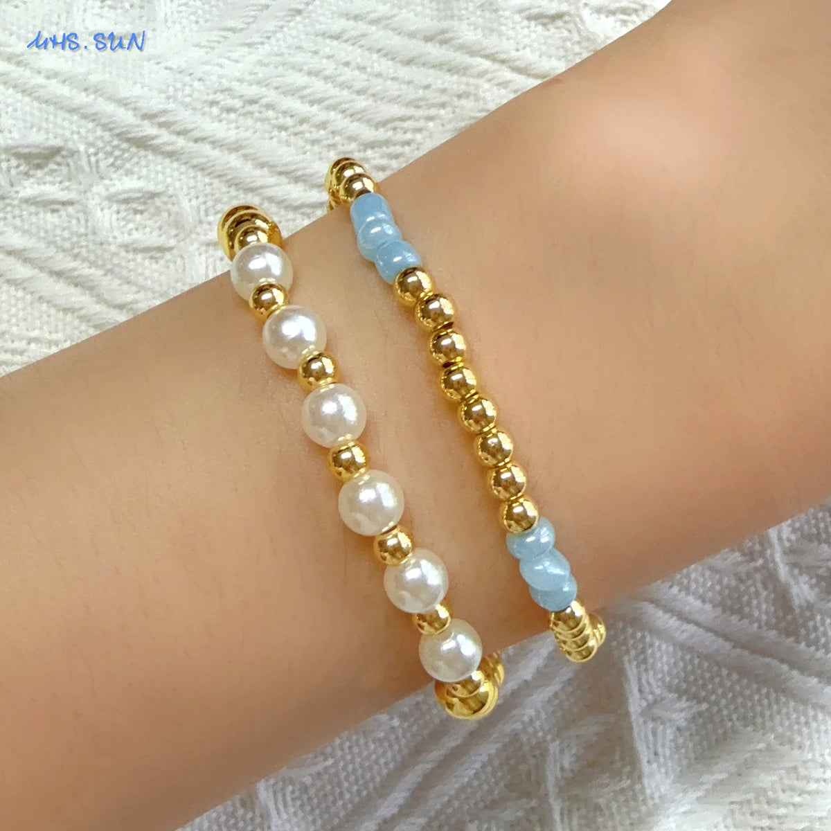 A gold-plated shell pearl beaded bracelet with white and light blue beads worn on a wrist.