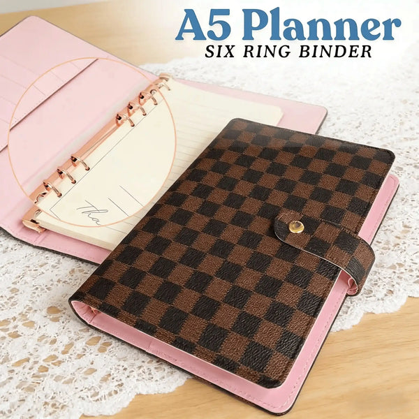 A pink A5 planner with a brown checkered cover and a leather binder shown on a wooden surface.