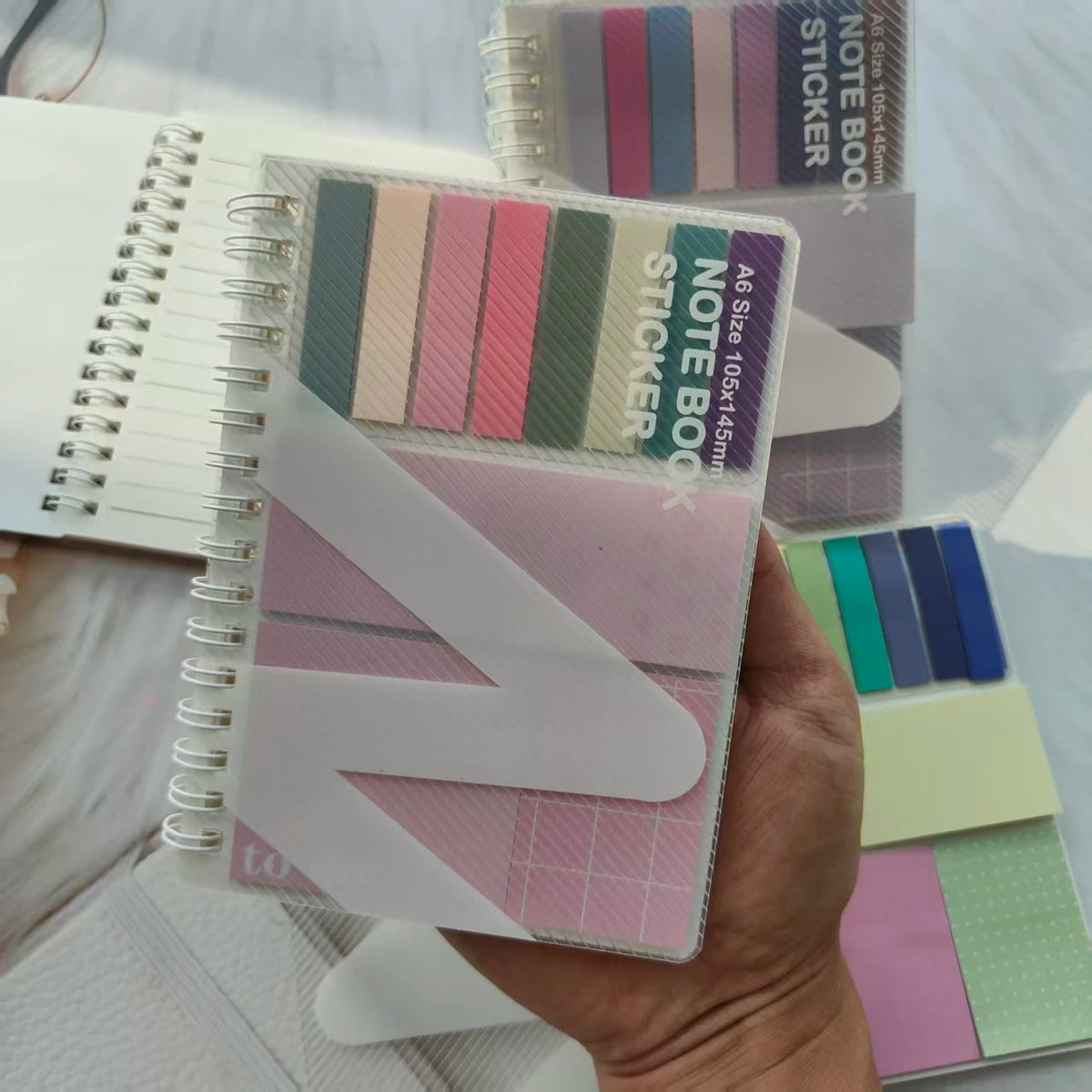 A hand holds an A6 sticky note notebook with pink and green colored sticky notes and a white page.