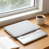 A silver laptop is placed on a wooden desk next to a gray PU leather sleeve, a notebook, and a white mug by a window.