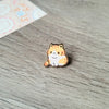Broche de esmalte con forma de gato atigrado naranja y blanco sentado sobre una superficie de madera.