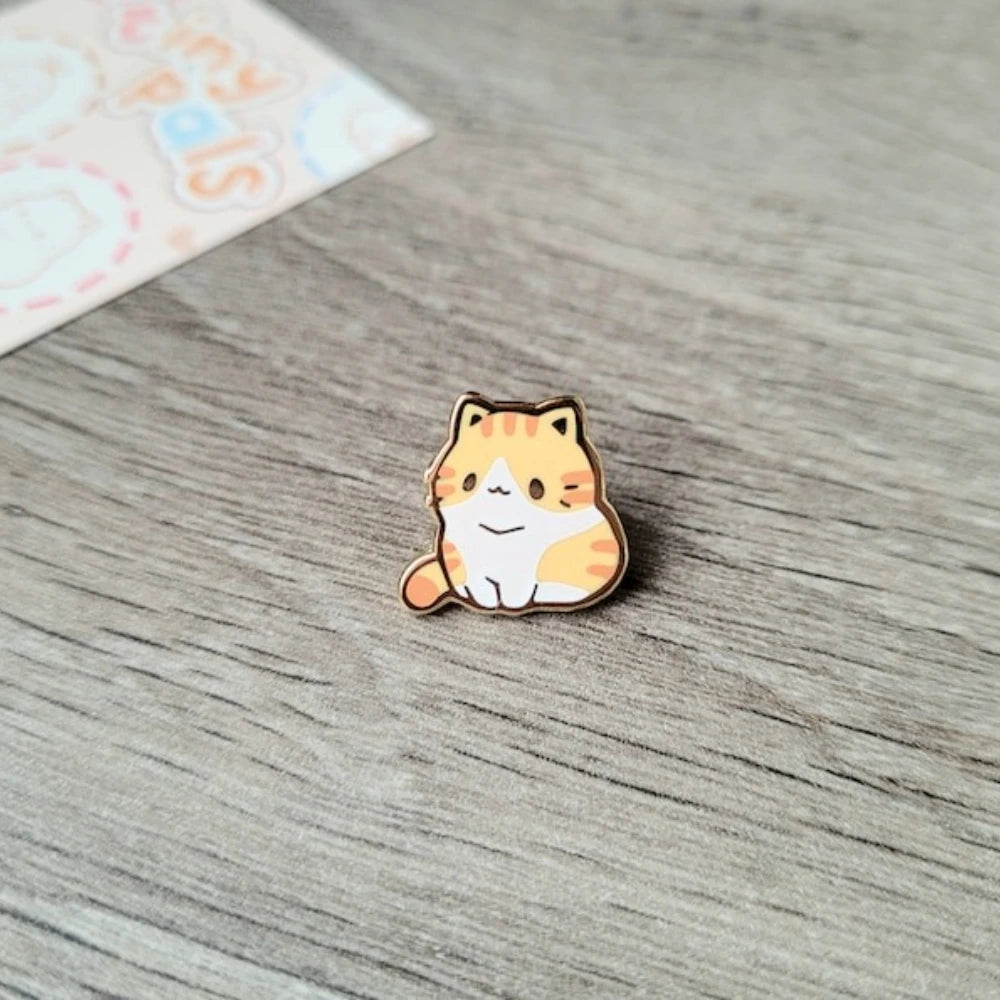 Broche de esmalte con forma de gato atigrado naranja y blanco sentado sobre una superficie de madera.