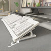 A white ergonomic keyboard riser stand with an anti-slip base is shown on a gray desk.