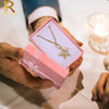 A person is holding a pink jewelry gift box with a gold necklace inside, set against a white background.