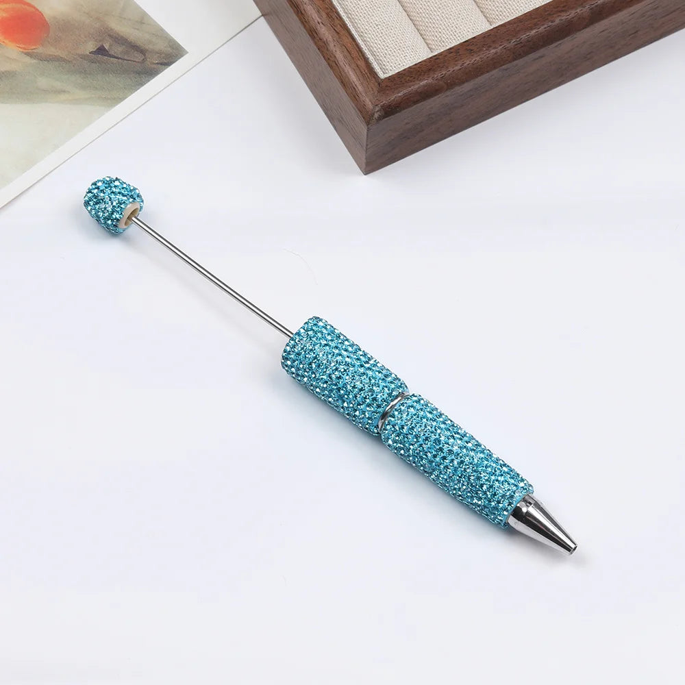 A silver pen accessory with a teal glitter ball and a blue glitter pen tip resting on a white surface.